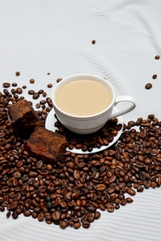 A white cup of coffee sits atop a saucer surrounded by a generous amount of roasted coffee beans. Next to the cup, there are two slices of chocolate cake. The textured white background contrasts with the rich, dark tones of the coffee beans and cake.