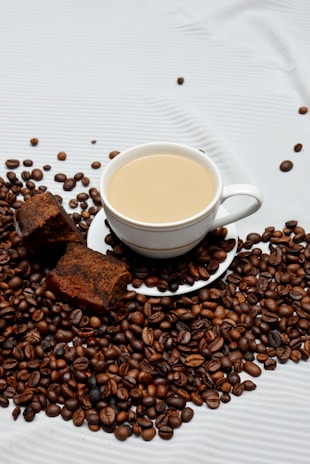 A white cup of coffee sits atop a saucer surrounded by a generous amount of roasted coffee beans. Next to the cup, there are two slices of chocolate cake. The textured white background contrasts with the rich, dark tones of the coffee beans and cake.