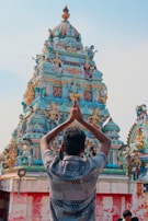 A person with folded hands is facing a colorful, intricately decorated Hindu temple. The temple is adorned with various statues and sculptures, showcasing detailed artistic craftsmanship.