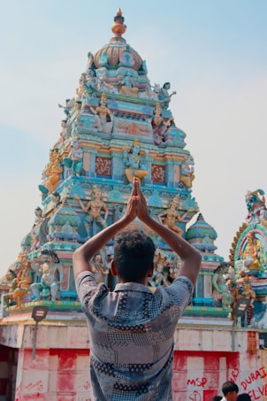 A person with folded hands is facing a colorful, intricately decorated Hindu temple. The temple is adorned with various statues and sculptures, showcasing detailed artistic craftsmanship.