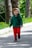 A young child is walking outdoors on a path surrounded by lush greenery. The child is wearing a dark green hoodie, bright red pants, and a cap, and appears happy and engaged with their surroundings. The background includes trees and a grassy area with small yellow flowers.