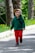 A young child is walking outdoors on a path surrounded by lush greenery. The child is wearing a dark green hoodie, bright red pants, and a cap, and appears happy and engaged with their surroundings. The background includes trees and a grassy area with small yellow flowers.