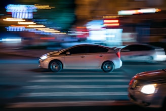 A car is moving quickly through a city street at night, with motion blur emphasizing speed. Bright lights in the background, both from nearby buildings and other vehicles, create a dynamic urban atmosphere.
