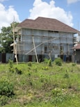 A house is under construction with scaffolding surrounding it. The building is two stories high and features a large, steeply pitched roof. The exterior walls are unfinished, and corrugated metal sheets form a temporary barrier around the site. The foreground shows a grassy area with scattered bushes, and the sky is partly cloudy.