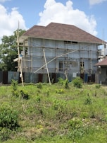 A house is under construction with scaffolding surrounding it. The building is two stories high and features a large, steeply pitched roof. The exterior walls are unfinished, and corrugated metal sheets form a temporary barrier around the site. The foreground shows a grassy area with scattered bushes, and the sky is partly cloudy.