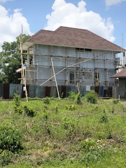 A house is under construction with scaffolding surrounding it. The building is two stories high and features a large, steeply pitched roof. The exterior walls are unfinished, and corrugated metal sheets form a temporary barrier around the site. The foreground shows a grassy area with scattered bushes, and the sky is partly cloudy.