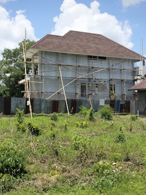 A house is under construction with scaffolding surrounding it. The building is two stories high and features a large, steeply pitched roof. The exterior walls are unfinished, and corrugated metal sheets form a temporary barrier around the site. The foreground shows a grassy area with scattered bushes, and the sky is partly cloudy.