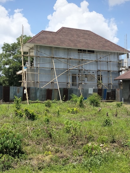 A house is under construction with scaffolding surrounding it. The building is two stories high and features a large, steeply pitched roof. The exterior walls are unfinished, and corrugated metal sheets form a temporary barrier around the site. The foreground shows a grassy area with scattered bushes, and the sky is partly cloudy.