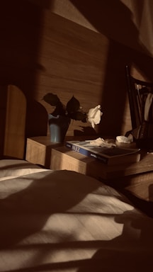 A cozy, softly lit bedroom corner with a nightstand holding a small vase of fresh flowers and a glowing lamp.