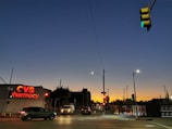 A city street scene at dusk with a dramatic gradient sky transitioning from deep blue to orange near the horizon. The intersection is illuminated by streetlights and traffic signals showing a green light. A CVS Pharmacy sign glows red on a nearby building. Vehicles, including a black car and a white van, are visible on the road. Utility poles and low-rise buildings are silhouetted against the evening sky.