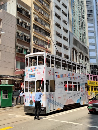 A person in a blue uniform is adjusting or maintaining a white tram with colorful advertisements. The tram is on a city street surrounded by tall buildings with many windows and air conditioning units. A red taxi is partially visible on the right side.
