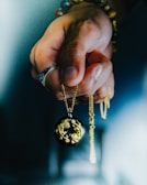 A hand holds a delicate pendant on a gold chain. The pendant is round and black with intricate gold lion designs. The person wears a silver ring and colorful beaded bracelets.