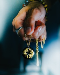 A hand holds a delicate pendant on a gold chain. The pendant is round and black with intricate gold lion designs. The person wears a silver ring and colorful beaded bracelets.