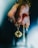 A hand holds a delicate pendant on a gold chain. The pendant is round and black with intricate gold lion designs. The person wears a silver ring and colorful beaded bracelets.
