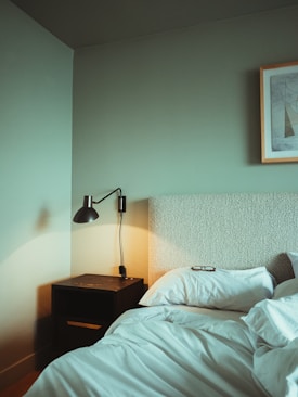 A minimalist bedroom features a bed with white sheets and a pillow on which a pair of glasses rests. Beside the bed is a dark wooden nightstand with a black modern lamp attached to the wall above it. The room has a soft green wall with a framed abstract artwork on it.