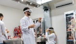 Several people wearing white lab coats engage with virtual reality headsets in a lab setting. Anatomical models and skeletons can be seen in the background, indicating an educational environment focused on anatomy. The individuals appear to be interacting with VR controllers, possibly simulating medical training or anatomical study.