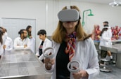 A woman wearing a virtual reality headset and holding controllers stands in a laboratory setting. She is dressed in a white lab coat and is surrounded by several people who are also in lab attire. Anatomical models are visible in the background, suggesting a medical or educational environment.