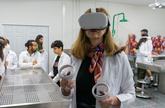 A woman wearing a virtual reality headset and holding controllers stands in a laboratory setting. She is dressed in a white lab coat and is surrounded by several people who are also in lab attire. Anatomical models are visible in the background, suggesting a medical or educational environment.