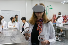 A woman wearing a virtual reality headset and holding controllers stands in a laboratory setting. She is dressed in a white lab coat and is surrounded by several people who are also in lab attire. Anatomical models are visible in the background, suggesting a medical or educational environment.