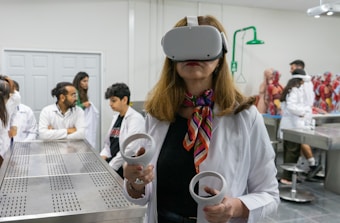A woman wearing a virtual reality headset and holding controllers stands in a laboratory setting. She is dressed in a white lab coat and is surrounded by several people who are also in lab attire. Anatomical models are visible in the background, suggesting a medical or educational environment.