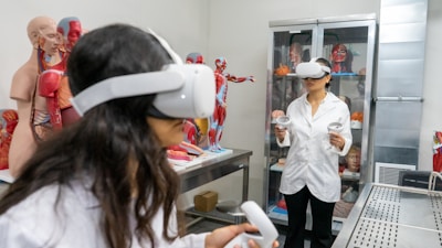 Two individuals wearing virtual reality headsets and holding controllers are inside a room filled with anatomical models and medical displays. Various anatomical figures, including muscles, organs, and skeletons, are visible. The individuals are dressed in lab coats, suggesting a scientific or educational setting.