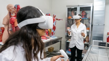 Two individuals wearing virtual reality headsets and holding controllers are inside a room filled with anatomical models and medical displays. Various anatomical figures, including muscles, organs, and skeletons, are visible. The individuals are dressed in lab coats, suggesting a scientific or educational setting.