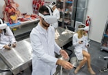Several individuals in white lab coats are wearing virtual reality headsets in a laboratory setting. Anatomical models and educational tools are visible on tables and shelves, indicating a medical or educational environment. The individuals appear to be using hand controllers while engaged in a virtual reality experience.