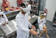 Several individuals in white lab coats are wearing virtual reality headsets in a laboratory setting. Anatomical models and educational tools are visible on tables and shelves, indicating a medical or educational environment. The individuals appear to be using hand controllers while engaged in a virtual reality experience.