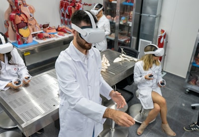 Several individuals in white lab coats are wearing virtual reality headsets in a laboratory setting. Anatomical models and educational tools are visible on tables and shelves, indicating a medical or educational environment. The individuals appear to be using hand controllers while engaged in a virtual reality experience.