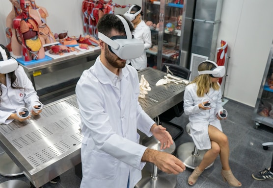Several individuals in white lab coats are wearing virtual reality headsets in a laboratory setting. Anatomical models and educational tools are visible on tables and shelves, indicating a medical or educational environment. The individuals appear to be using hand controllers while engaged in a virtual reality experience.