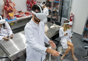 Several individuals in white lab coats are wearing virtual reality headsets in a laboratory setting. Anatomical models and educational tools are visible on tables and shelves, indicating a medical or educational environment. The individuals appear to be using hand controllers while engaged in a virtual reality experience.