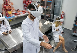 Several individuals in white lab coats are wearing virtual reality headsets in a laboratory setting. Anatomical models and educational tools are visible on tables and shelves, indicating a medical or educational environment. The individuals appear to be using hand controllers while engaged in a virtual reality experience.
