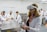 A woman is wearing a virtual reality headset and holding controllers in her hands. She is in a laboratory setting with several other people, all wearing lab coats, sitting at a stainless steel table. The background shows more individuals engaged in activities, some interacting with anatomical models.