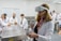 A woman is wearing a virtual reality headset and holding controllers in her hands. She is in a laboratory setting with several other people, all wearing lab coats, sitting at a stainless steel table. The background shows more individuals engaged in activities, some interacting with anatomical models.