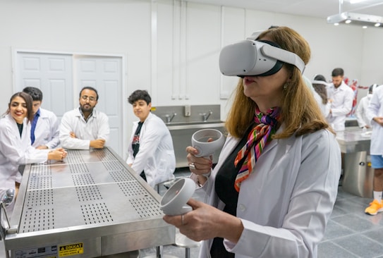A woman is wearing a virtual reality headset and holding controllers in her hands. She is in a laboratory setting with several other people, all wearing lab coats, sitting at a stainless steel table. The background shows more individuals engaged in activities, some interacting with anatomical models.