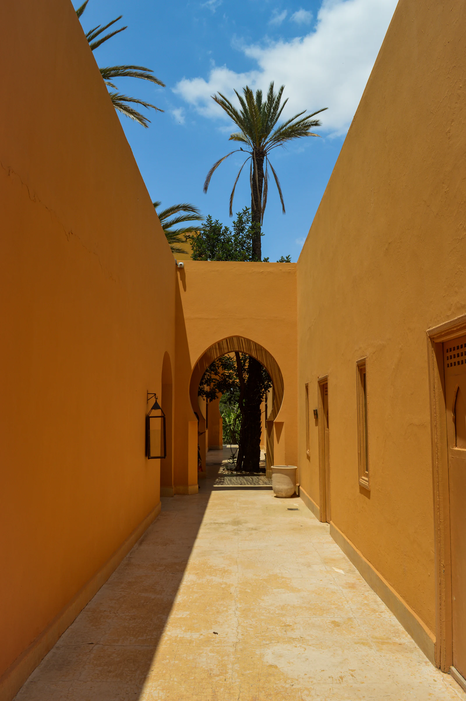 Allée entre des murs ocre, arche et palmiers sous un ciel bleu, à Marrakech
