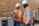 Two individuals wearing white hard hats and orange safety vests are in a workshop or industrial setting. One person is holding a concrete block while the other observes. They are standing next to a machine with a brick wall in the background.