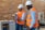 Two individuals wearing white hard hats and orange safety vests are in a workshop or industrial setting. One person is holding a concrete block while the other observes. They are standing next to a machine with a brick wall in the background.