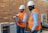 Two individuals wearing white hard hats and orange safety vests are in a workshop or industrial setting. One person is holding a concrete block while the other observes. They are standing next to a machine with a brick wall in the background.