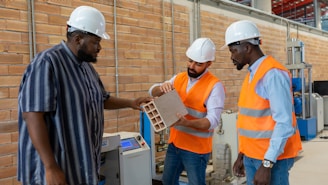 Three individuals wearing safety helmets and orange vests are engaged in a discussion about a construction material inside a workshop or factory setting. The environment is industrial, with visible machinery and brick walls in the background. One person is holding a rectangular block, possibly a type of building material, while the others observe and interact.