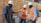 Three individuals wearing safety helmets and orange vests are engaged in a discussion about a construction material inside a workshop or factory setting. The environment is industrial, with visible machinery and brick walls in the background. One person is holding a rectangular block, possibly a type of building material, while the others observe and interact.