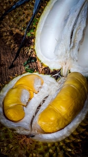 A close-up view of an open durian fruit with vibrant yellow flesh inside. The spiky outer shell is visible, along with the creamy texture of the fruit's interior.