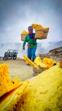 A person is carrying baskets filled with bright yellow sulfur. The background displays a smoky, mountainous terrain, possibly a volcanic area. In the forefront are large chunks of yellow sulfur, and an empty cart is visible on the side.