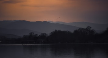 A serene landscape featuring a calm body of water in the foreground, with reflections of silhouetted trees along the shoreline. The background is dominated by layers of rolling hills and mountains, creating a sense of depth. The sky above transitions from a warm orange hue to a darker tone near the top, suggesting early morning or evening lighting. A few birds are visible in the distance, adding a sense of life to the tranquil scene.
