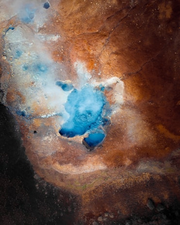 An aerial view of a geothermal area featuring a vivid blue pool surrounded by various shades of brown, rust, and orange earth. Steam is rising from the pool, indicating thermal activity. The textures of the ground appear rugged and uneven.