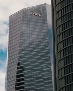 A modern high-rise building with reflective glass windows and the KPMG logo near the top. The structure features sleek, vertical lines and a mix of metal and glass materials. Another building with a curved fa&ccedil;ade is partially visible on the right side.