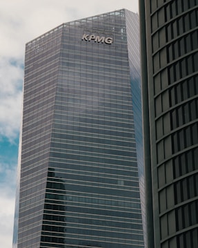 A modern high-rise building with reflective glass windows and the KPMG logo near the top. The structure features sleek, vertical lines and a mix of metal and glass materials. Another building with a curved fa&ccedil;ade is partially visible on the right side.