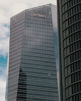 A modern high-rise building with reflective glass windows and the KPMG logo near the top. The structure features sleek, vertical lines and a mix of metal and glass materials. Another building with a curved façade is partially visible on the right side.