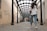 A long, covered walkway with a glass roof and black metal supports leads into the distance. The walkway is lined with light-colored walls, some of which are covered with graffiti. A young woman in a white hoodie and jeans walks away from the camera towards a group of people in the distance.