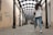 A long, covered walkway with a glass roof and black metal supports leads into the distance. The walkway is lined with light-colored walls, some of which are covered with graffiti. A young woman in a white hoodie and jeans walks away from the camera towards a group of people in the distance.